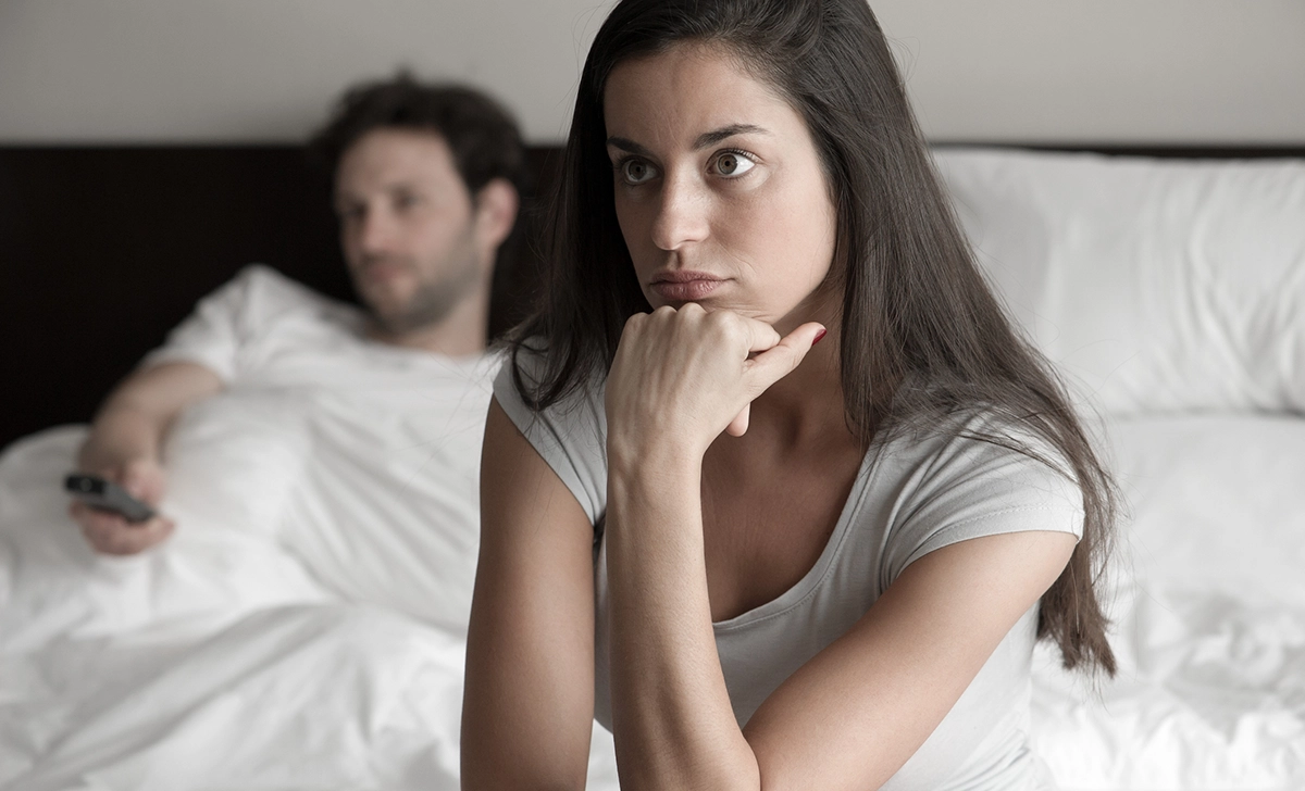 A woman sits on the edge of a bed with a pensive, unhappy expression, resting her chin on her hand. Behind her, a man lies back on the bed, holding a remote and looking away. Their body language suggests emotional distance and disconnection, reflecting tension and possible thoughts of divorce.