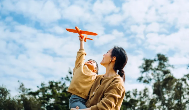 Parent holding a child as they play with a toy airplane outdoors.