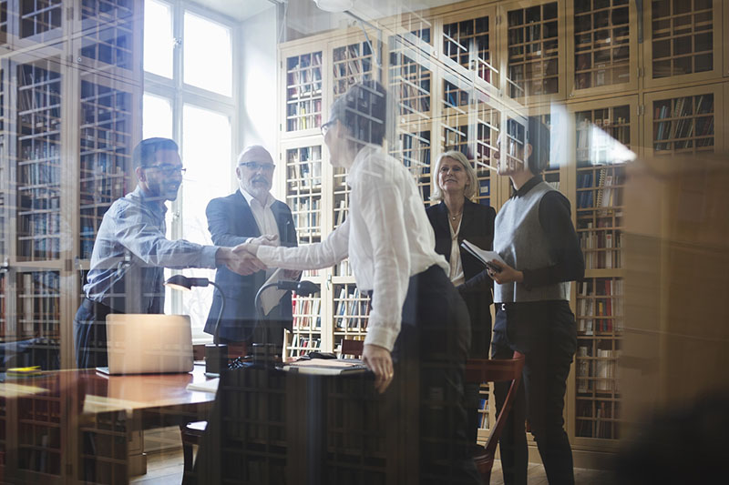 Business professionals shaking hands in a modern office library setting, viewed through a glass wall, symbolizing collaboration and agreement.