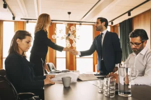 Female attorney and male attorney shake hands across a conference table while colleagues review legal documents during a divorce-settlement meeting.