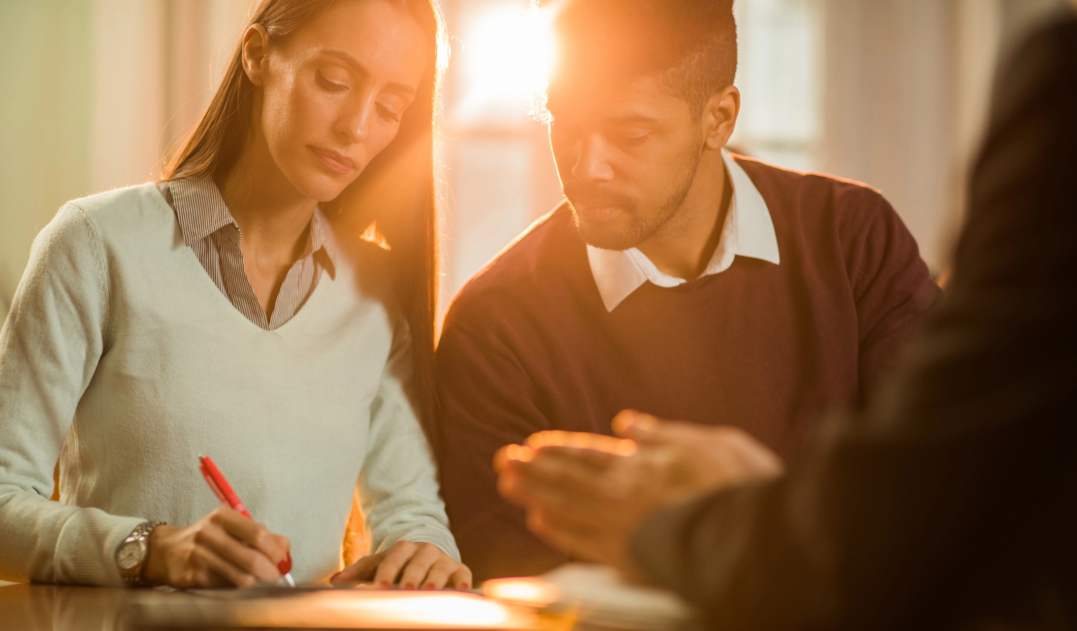 A couple sits together at a desk during a meeting, with the woman signing a document and the man looking on attentively. They appear calm and focused, while a third person, likely a mediator or lawyer, gestures from across the table. The warm lighting conveys a cooperative and respectful atmosphere, symbolizing a collaborative divorce process.