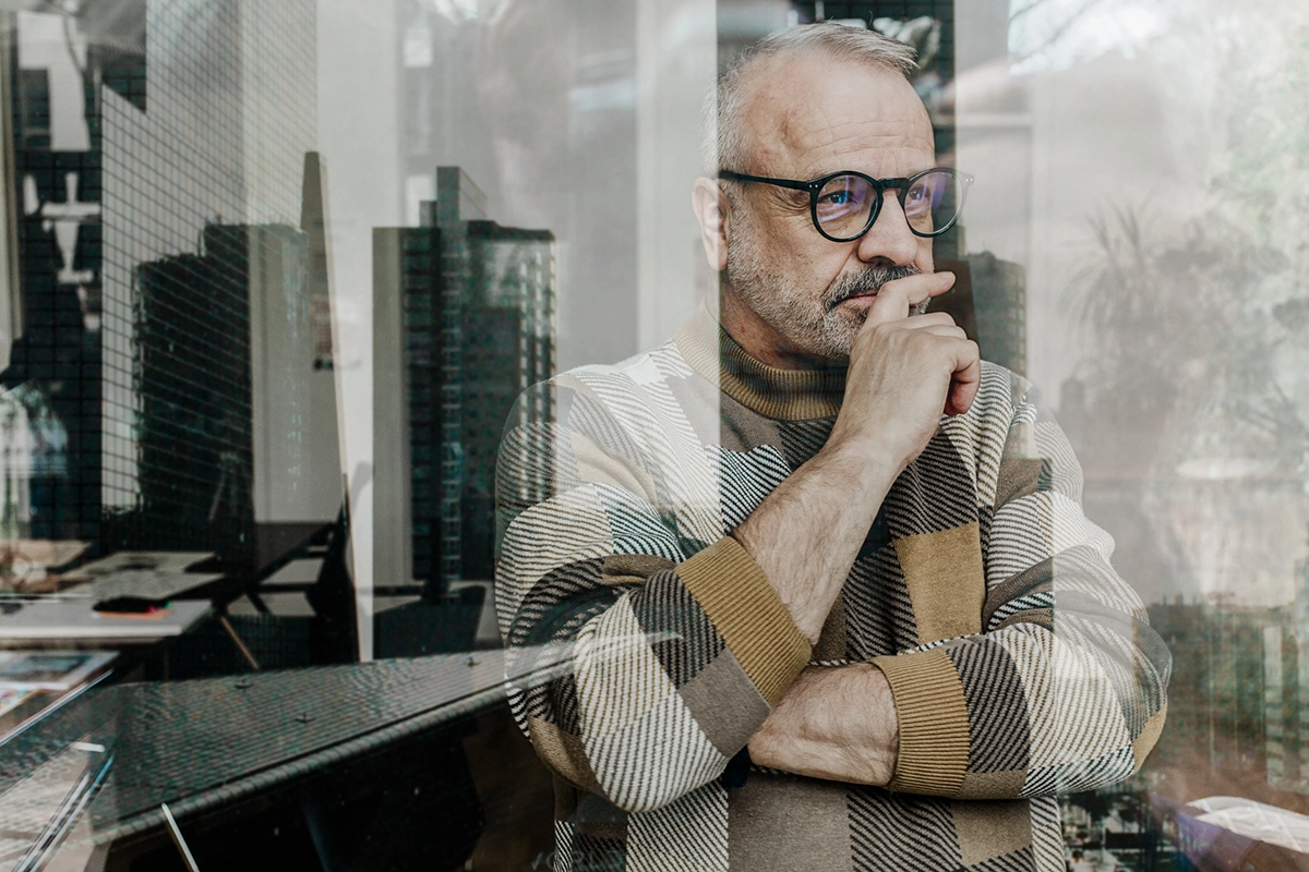 An older man with glasses stands indoors, looking thoughtfully out of a window. His hand rests near his mouth as he gazes into the distance. Reflections of tall city buildings appear in the glass in front of him, blending with the interior behind him. He wears a patterned sweater and appears deep in contemplation.