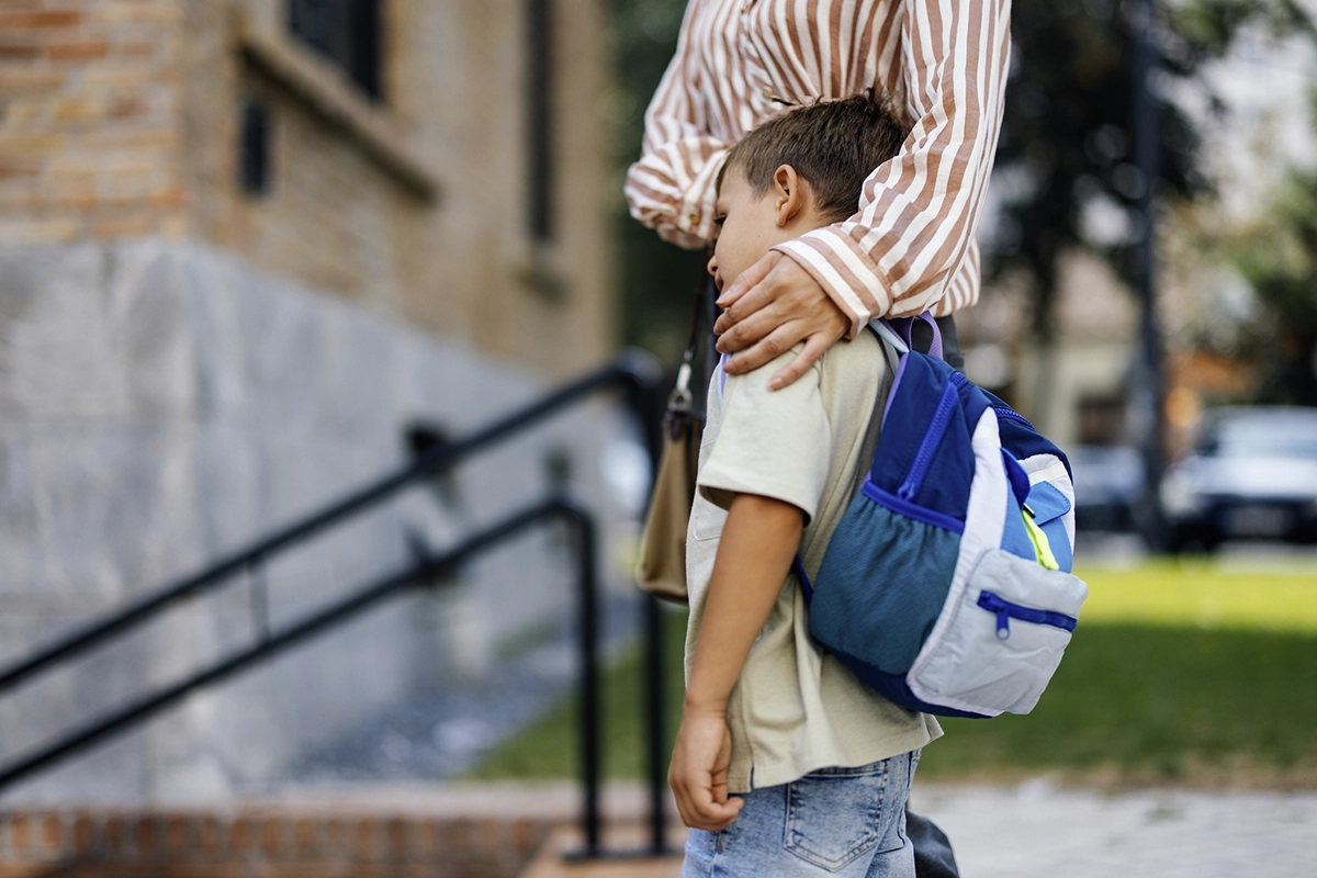 Young child wearing a backpack standing outdoors near a building, while an adult places a comforting hand on the child’s shoulder, suggesting reassurance or support before school.