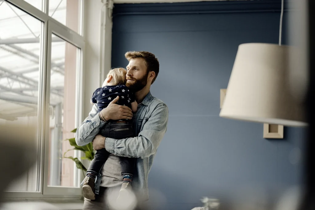 Man standing indoors holding a small child against his chest near a window, gently cradling the child in a calm, softly lit living space.