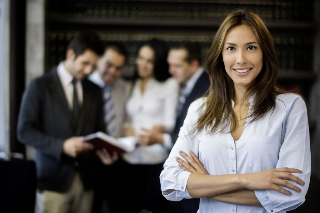 Confident professional woman standing with arms crossed and smiling in an office setting, with a group of colleagues blurred in the background reviewing documents.