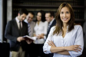 Confident professional woman standing with arms crossed and smiling in an office setting, with a group of colleagues blurred in the background reviewing documents.