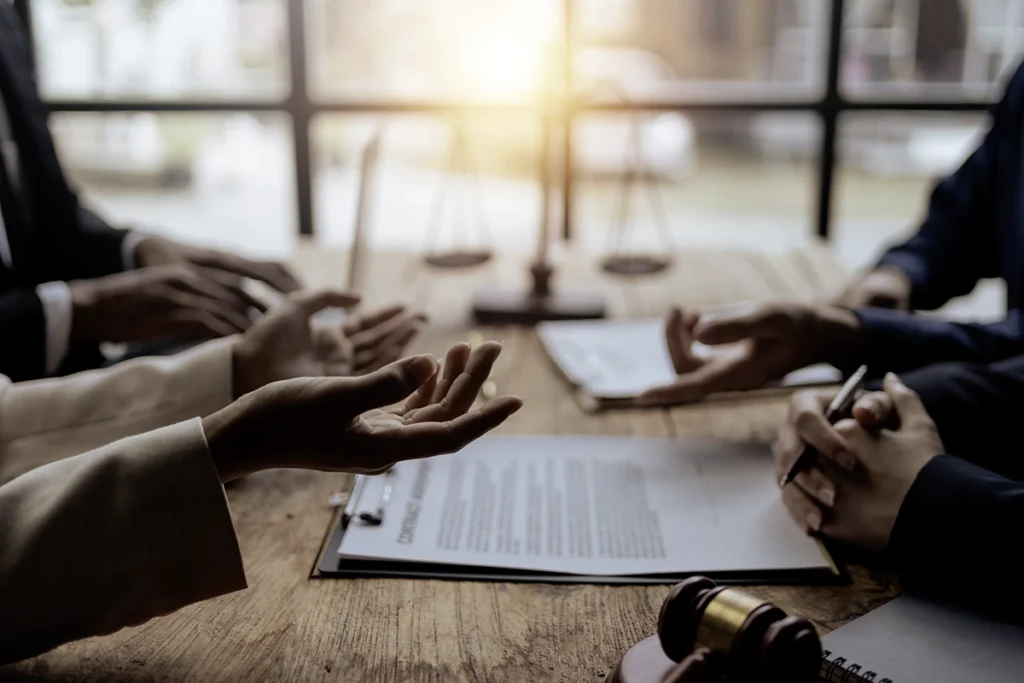 Close-up of hands gesturing during a legal discussion across a table with documents, a gavel, and scales of justice visible in a professional office setting.