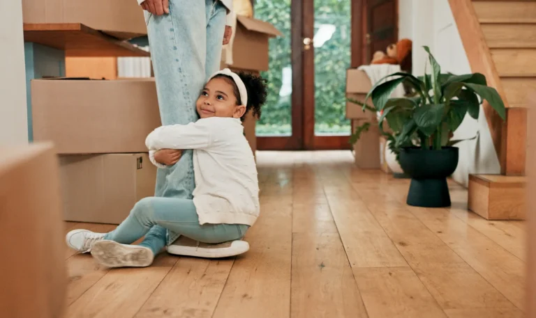 Child hugging a parent’s leg while sitting on the floor in a home with moving boxes.
